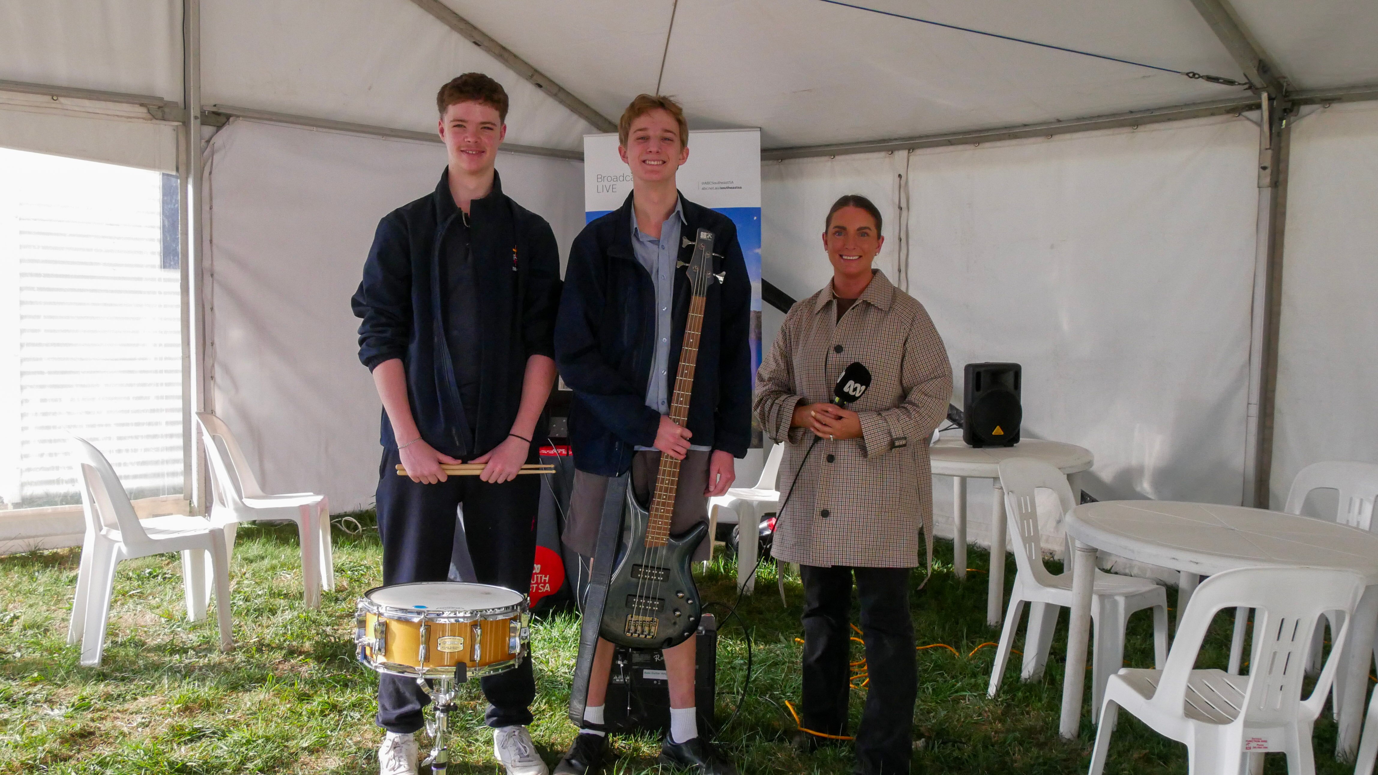 Three people in an outside area, two with instruments, one is a radio presenter with a microphone.