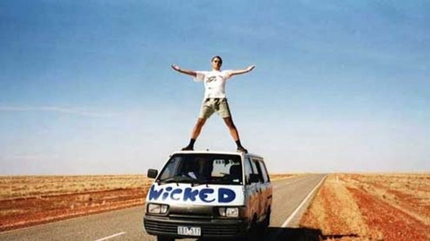 A man stands on top of a Wicked campervan with desert in background.