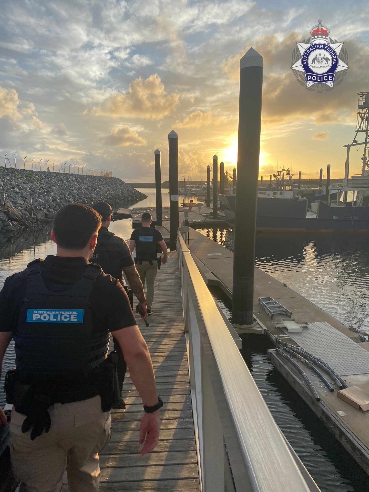 Tres policías caminan por un muelle hacia un grupo de barcos amarrados. 