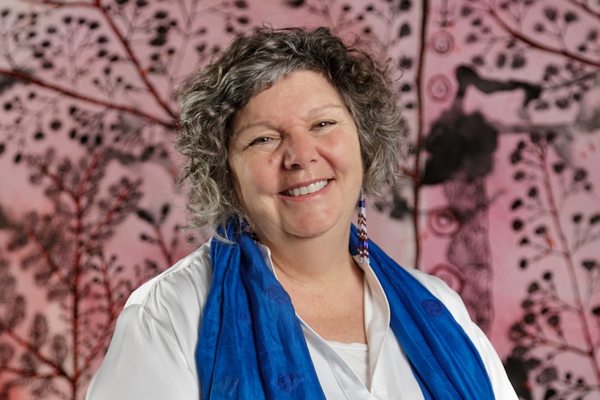 Woman with short grey curly hair wearing white shirt and blue scarf smiling, with pink and black artwork behind.