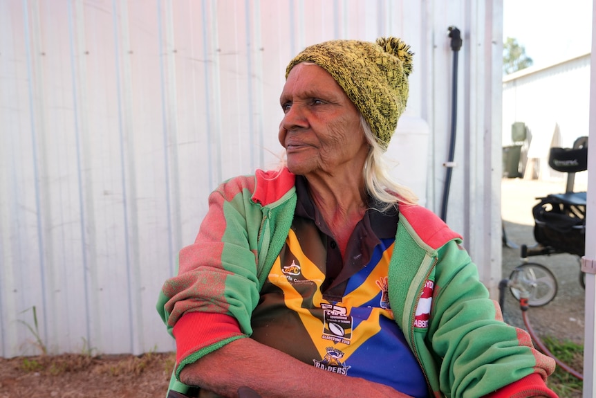 A woman sits in the shade in front of a shed and looks to her right into the distance.