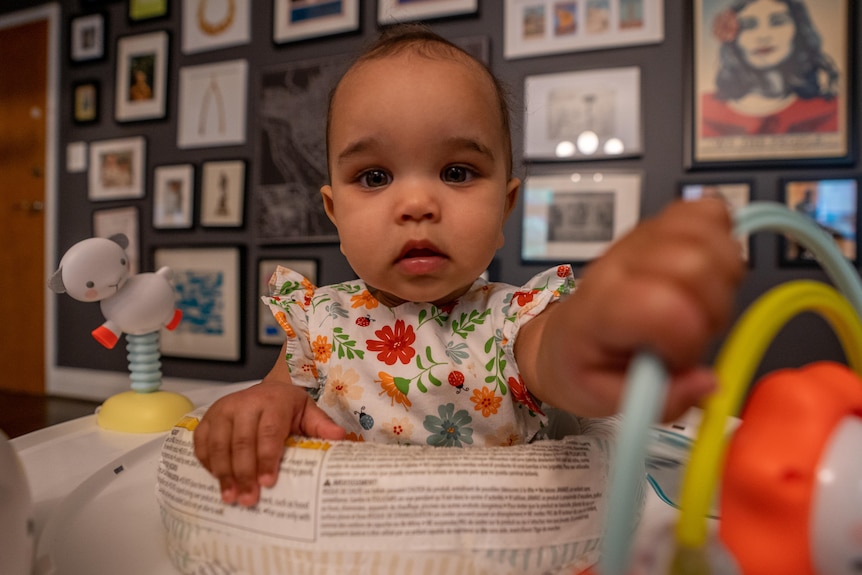 A baby in a floral jumpsuit holds a rattle