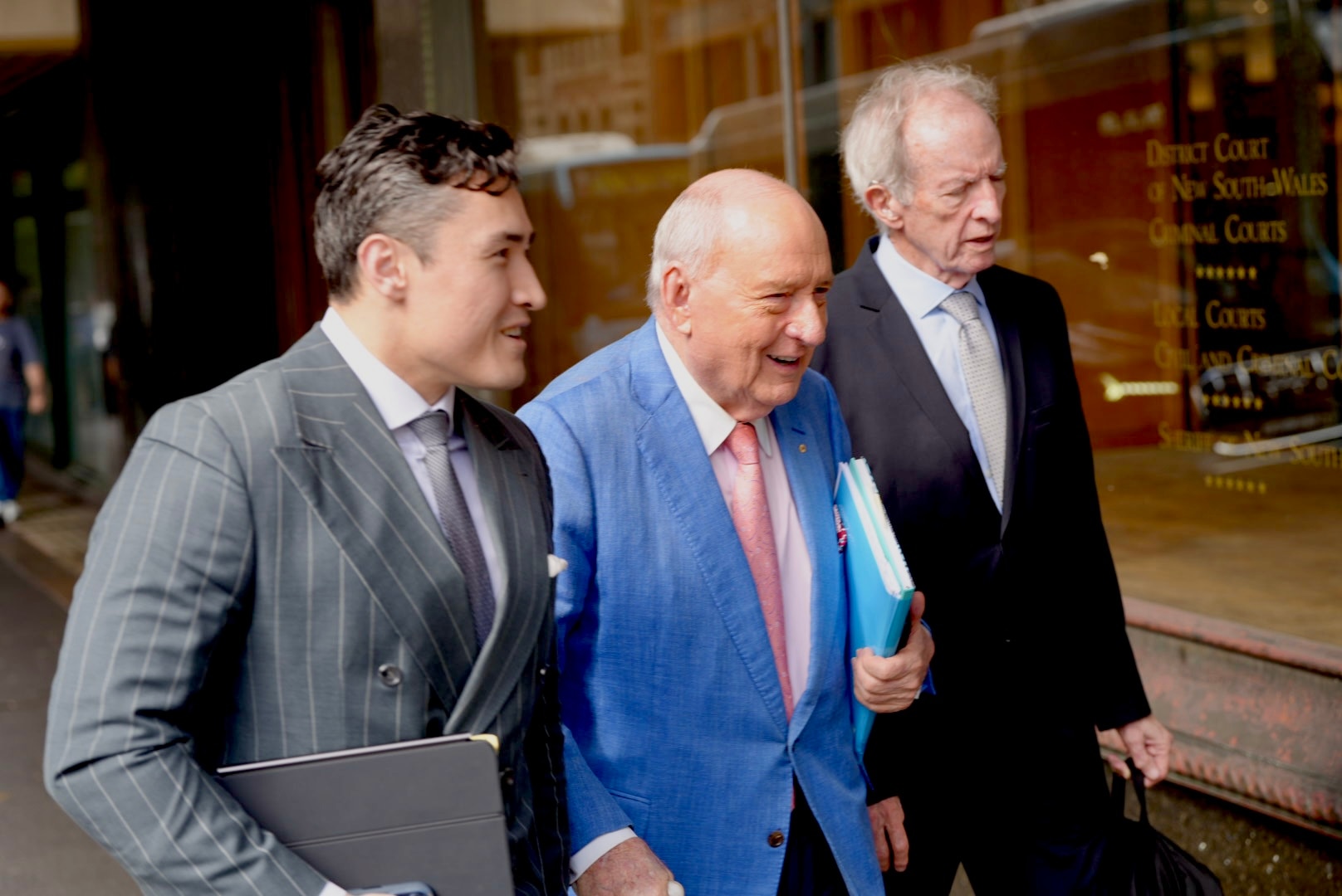 Man in blue jacket, pink tie and walking stick walks with two men in suits either side of him