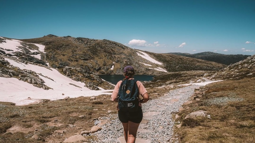 A woman walking along a hiking track, with mountains and a lake nearby.