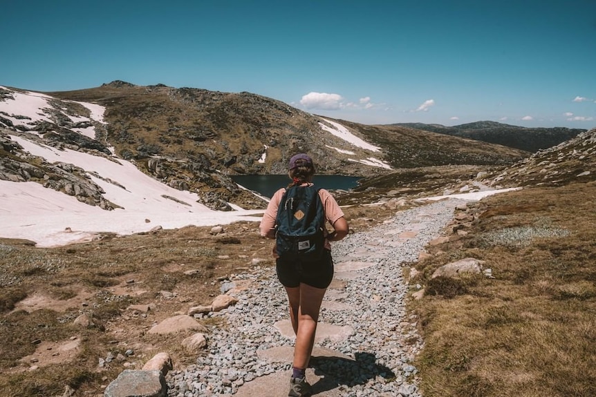 A woman walking along a hiking track, with mountains and a lake nearby.