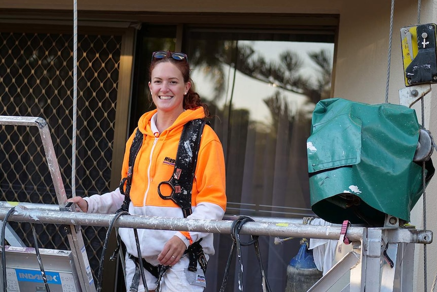 Woman with bright orange and white jumper and safety gear strapped to her on a swing stage outside someone's balcony