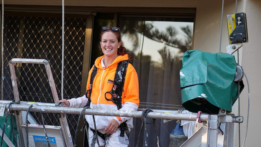 Woman with bright orange and white jumper and safety gear strapped to her on a swing stage outside someone's balcony