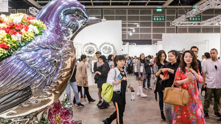 Chinese people visiting an art gallery walk past a large silver sculpture of a bird with flowers on its back.