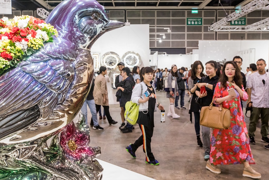 Chinese people visiting an art gallery walk past a large silver sculpture of a bird with flowers on its back.