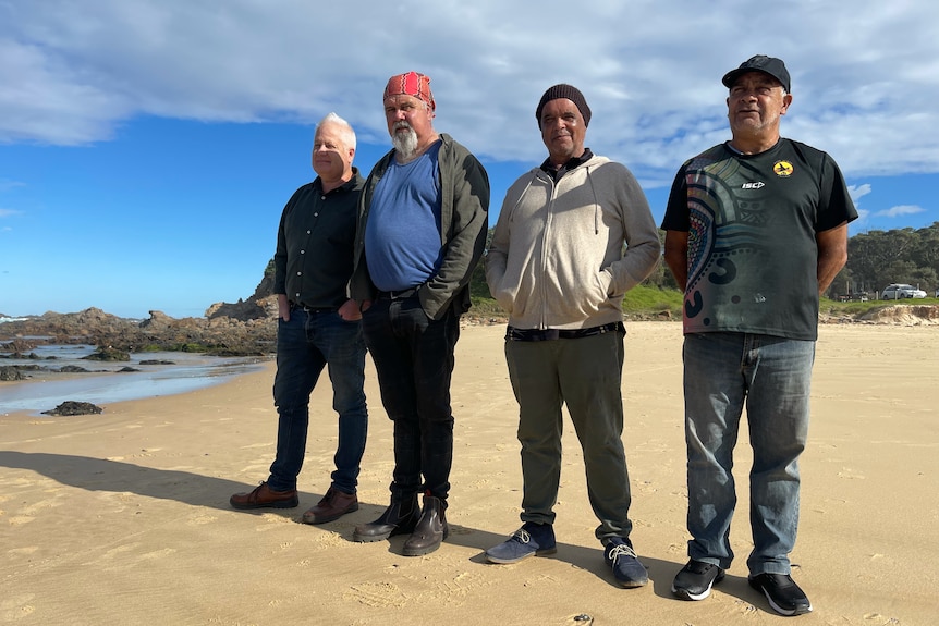 Four Yuin men stand in a row on the beach, looking serious.