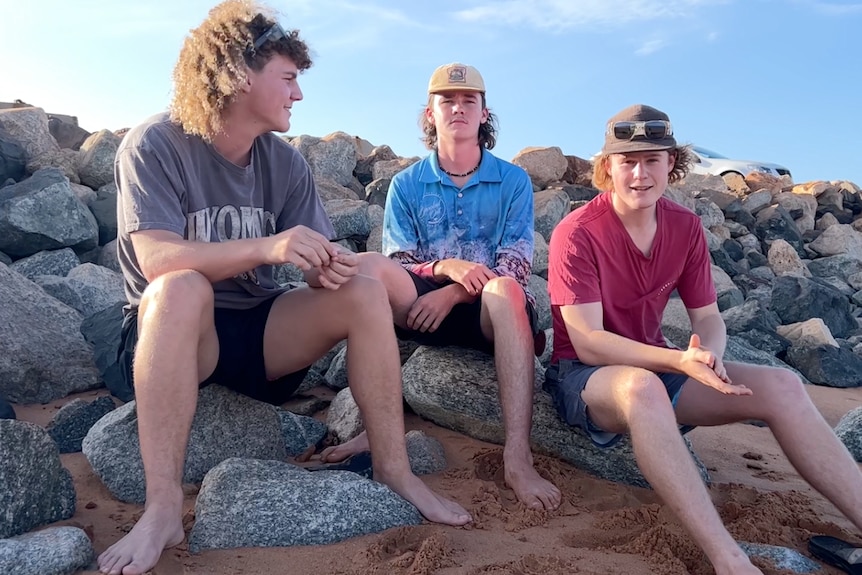 Three teenage boys sit on rocks at beach, one has long curly hair, other two wear caps, all wear shorts, smile.