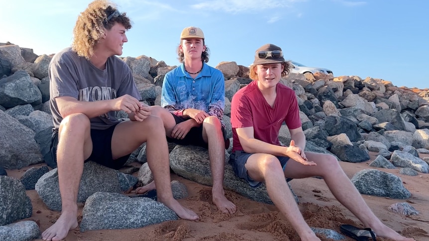 three teenage boys sit on rocks at beach
