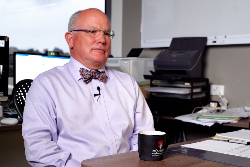 Man in a button-up shirt and bow tie sits at a desk. He is bald, wearing glasses, and facing us but looking to the right.