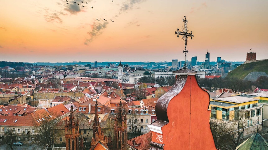 A skyline of Vilnius at dusk shows a cluster of medieval, orange rooftops, from behind a church spire.