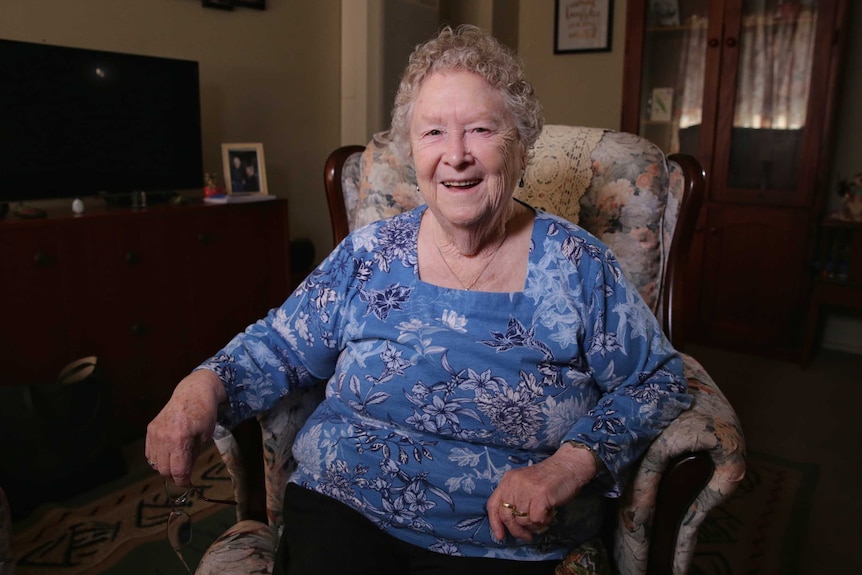 An elderly woman sits in an ornate armchair, with a sparkle in her eye and smile on her face. She is wearing a blue floral top.