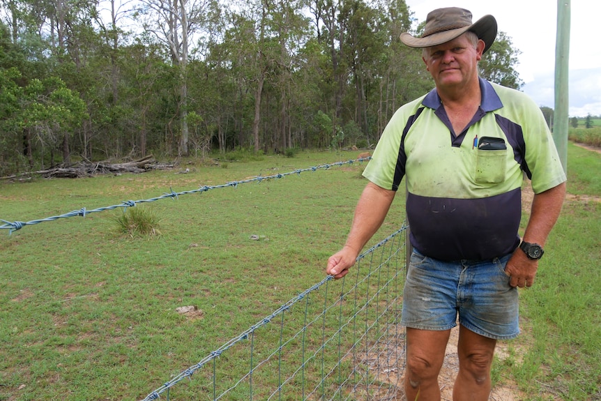 An older man in  tatty work clothes and a brown wide brimmed hat holds a pig proof fence separating his farm from his neighbours