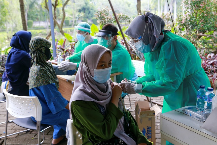 Three Asian women in Islamic headscarves get a COVID jab from nurses in full PPE, at an outside pop up clinic.