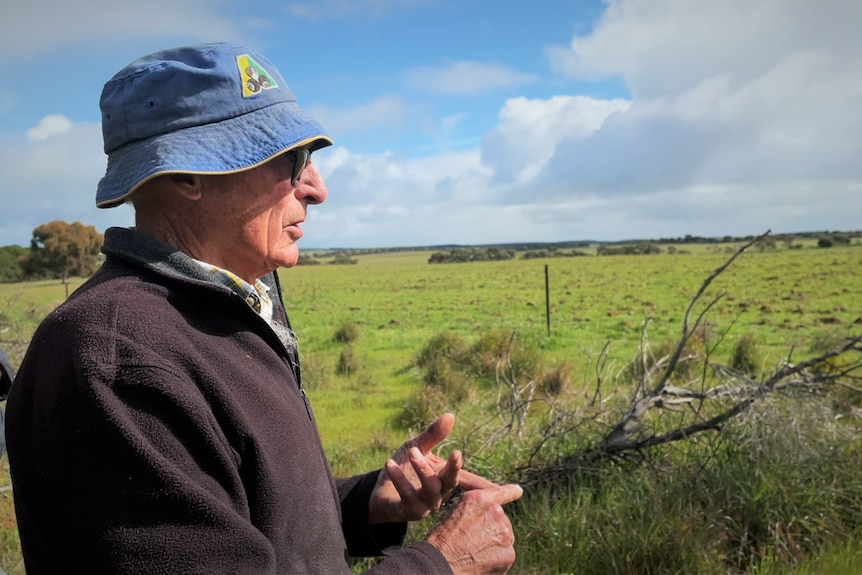 A man wears sunglasses, a blue hat and a dark jumper looks over green farming land