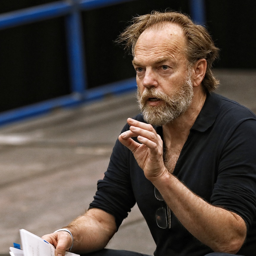 Hugo Weaving, sitting on the edge of a stage, reads from a script.