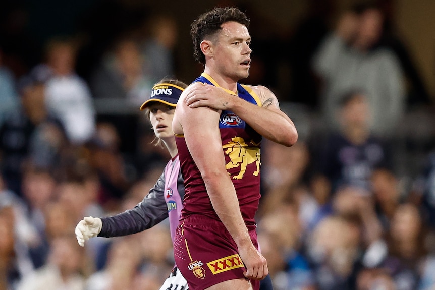 Brisbane Lions player Lachie Neale stands on the ground holding his sore shoulder as a team trainer watches. 