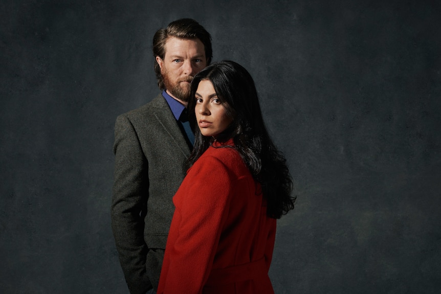 Man in suit stands behind woman in red coat, both staring at camera.