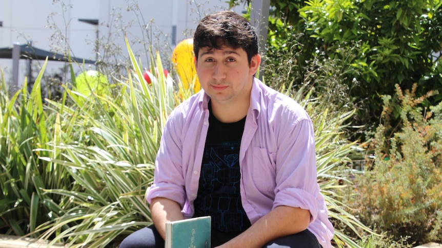 Andre May-Dessmann holding a book and sitting on a wall with plants in the background.