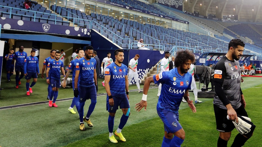 Football players wearing blue shirts walk out into an empty stadium