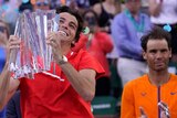 Tennis player Taylor Fritz lifts a large crystal trophy while smiling, as Rafael Nadal applauds behind him.