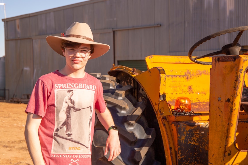 A white male teenager wearing an Akubra standing next to a yellow rusted tractor. He has a neutral expression.