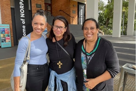 Three women stand close together with their arms around each other and smiling