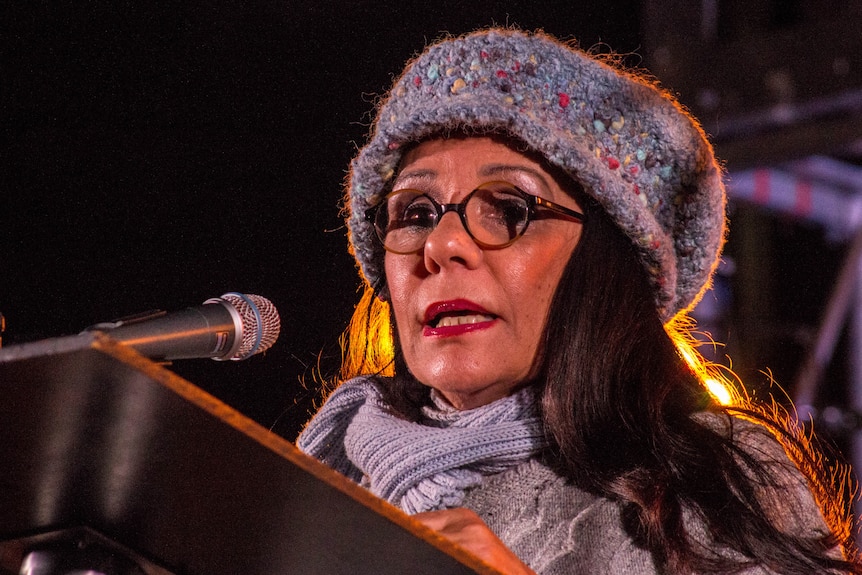 A woman wearing a winter hat, scarf and jumper speaks to a crowd of people on a June Winter's night.