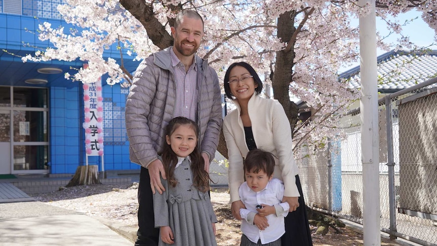 Parents and two kids smiling at the camera in Japan, with a blooming cherry blossom tree in the background.