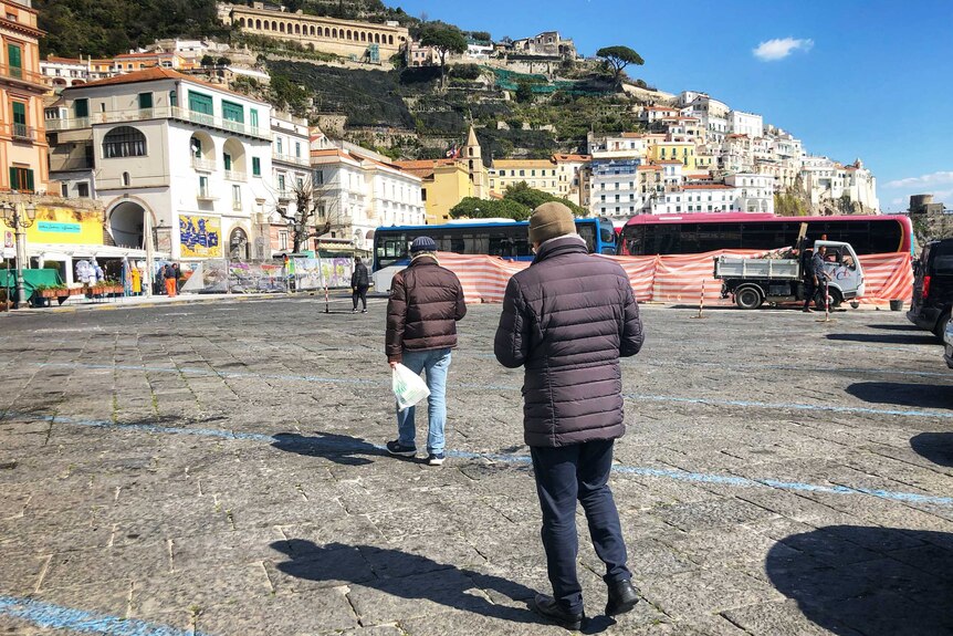 Two men in puffer jackets walking along a town square in Amalfi, Italy