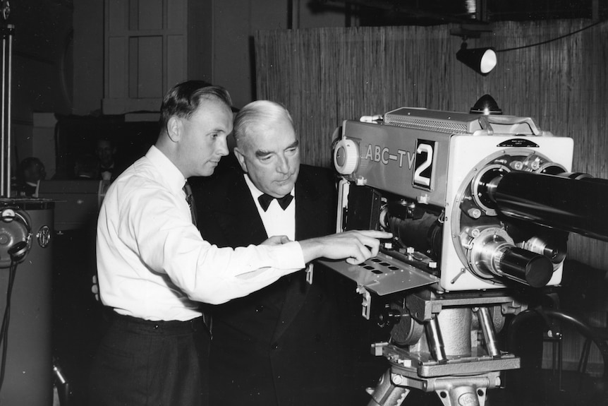 Black and white photo of two men looking at an old fashioned film camera.
