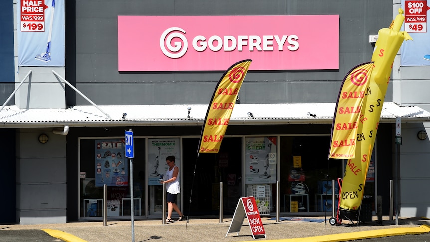 A person leaves a Godfreys retail store with large "sale" banners out the front.