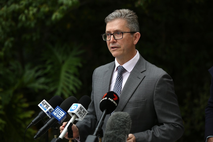 A man in a grey suit stands in front of a bank of microphones. 