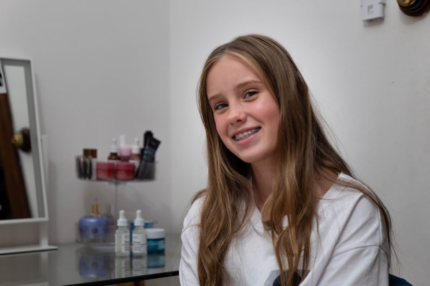 A smiling young Caucasian girl with braces and long blond hair, sitting near a mirror and skincare products on a desk.