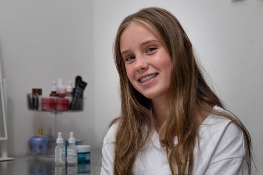 A smiling young Caucasian girl with braces and long blond hair, sitting near a mirror and skincare products on a desk.