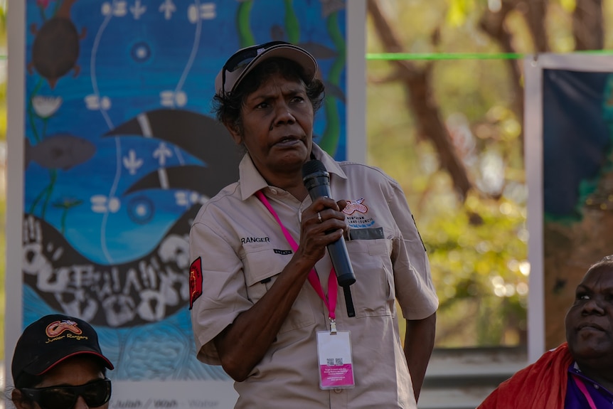 An Aboriginal woman holds a microphone and addressed a crowd. She looks serious.