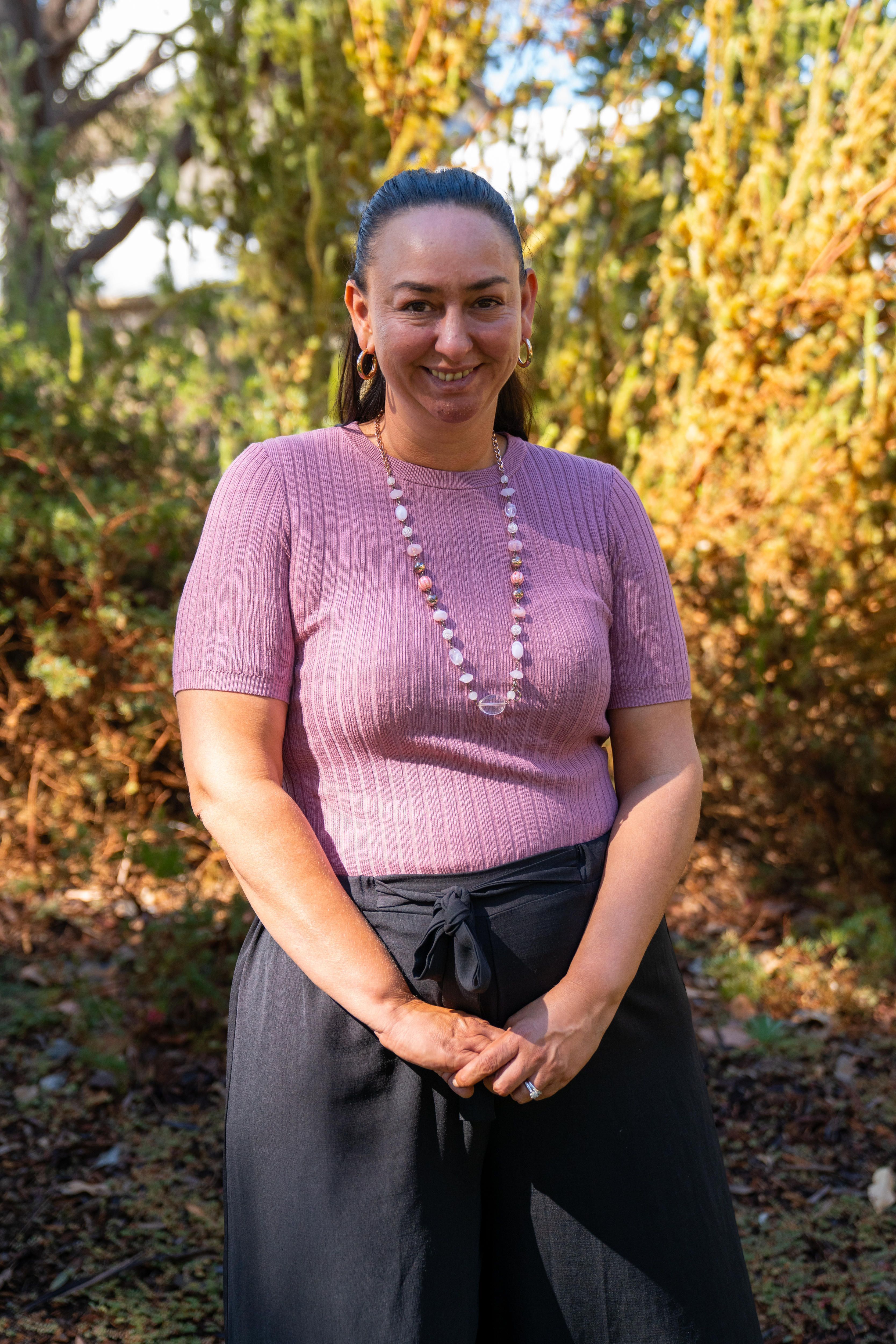 A woman wearing a pink shirt, long white-and-pink necklace and black pants smiles. She's in front of some trees, in a garden.