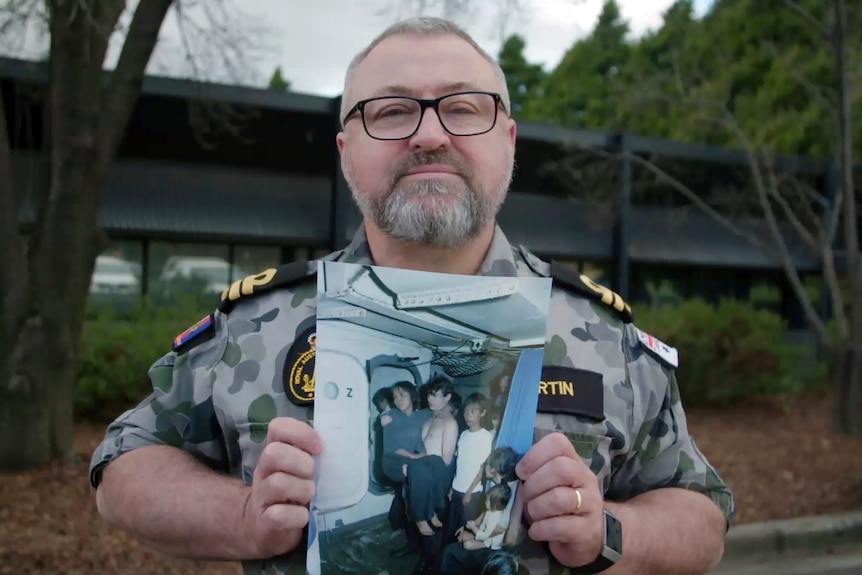 A man wearing glasses and a Navy uniform holds a photo of his younger self carrying a refugee aboard a war ship