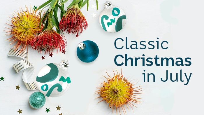 Text: 'Classic Christmas in July', set amongst native Australian flowers and blue baubles