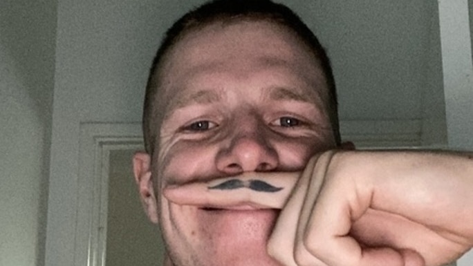 A shirtless man with short hair holds his finger above his lip, and on his finger is a tattoo of a moustache