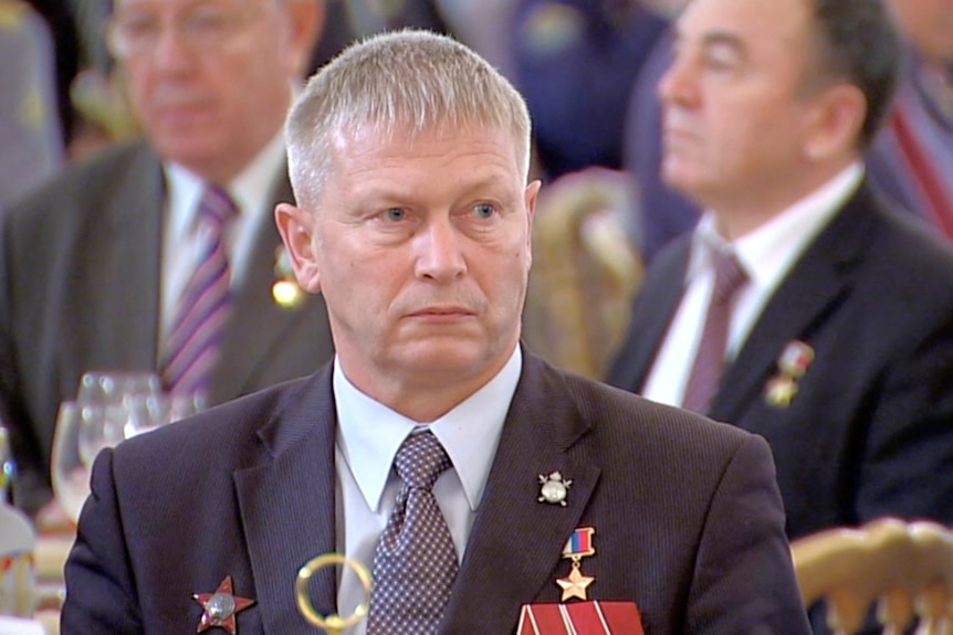 An older man with white hair sits in a suit with military medals on the lapel