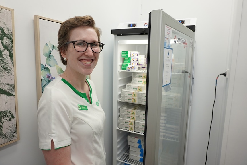 Woman in pharmacist uniform stands next to vaccine fridge smiling.