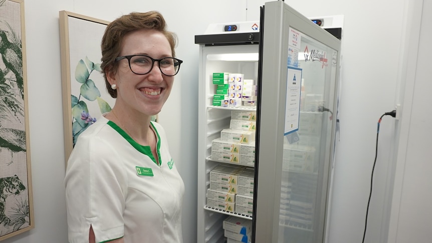 Woman in pharmacist uniform stands next to vaccine fridge smiling.