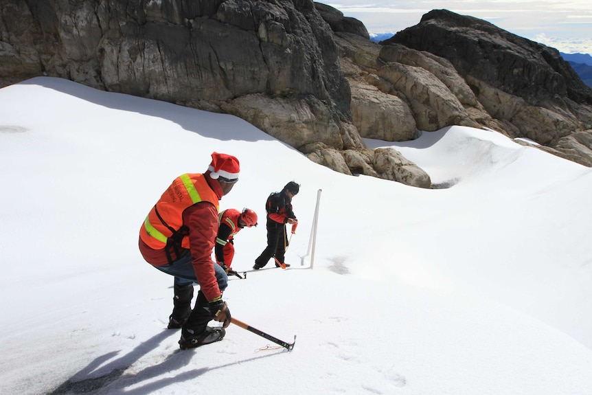 Three people, one wearing a santa hat, stand on sloping ice on a mountain. One is setting up equipment.