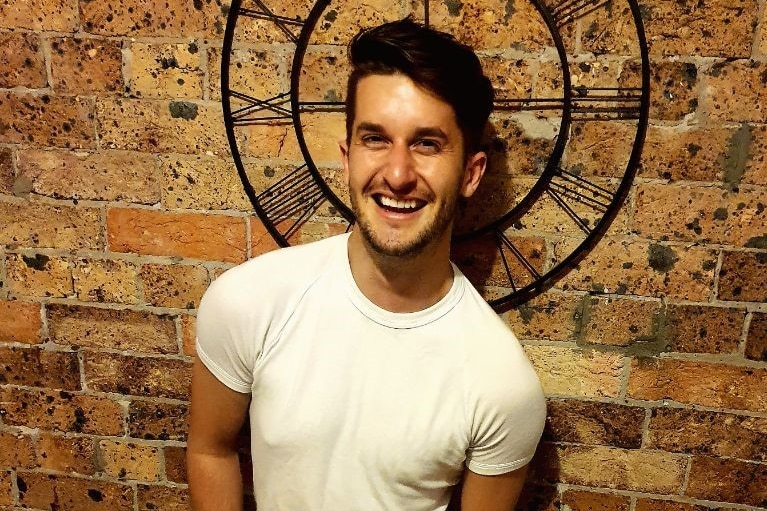 A young man stands laughing in front of a clock on a brick wall, wearing a white t-shirt and grey pants.