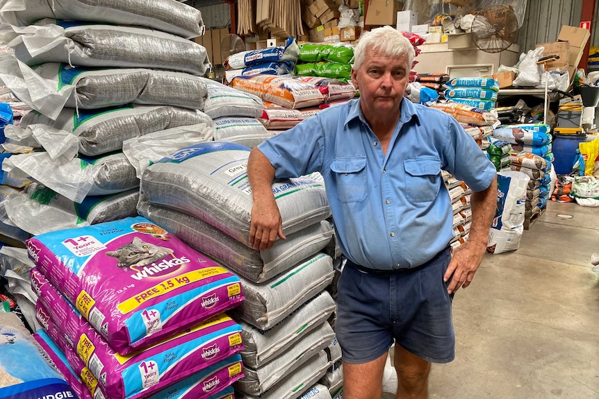 A man with grey hair, blue shirt, leaning against bags of animal feed in a shed.