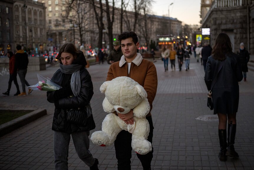 A couple holding a big stuffed teddy bear and a rose walk through a square in Kyiv.